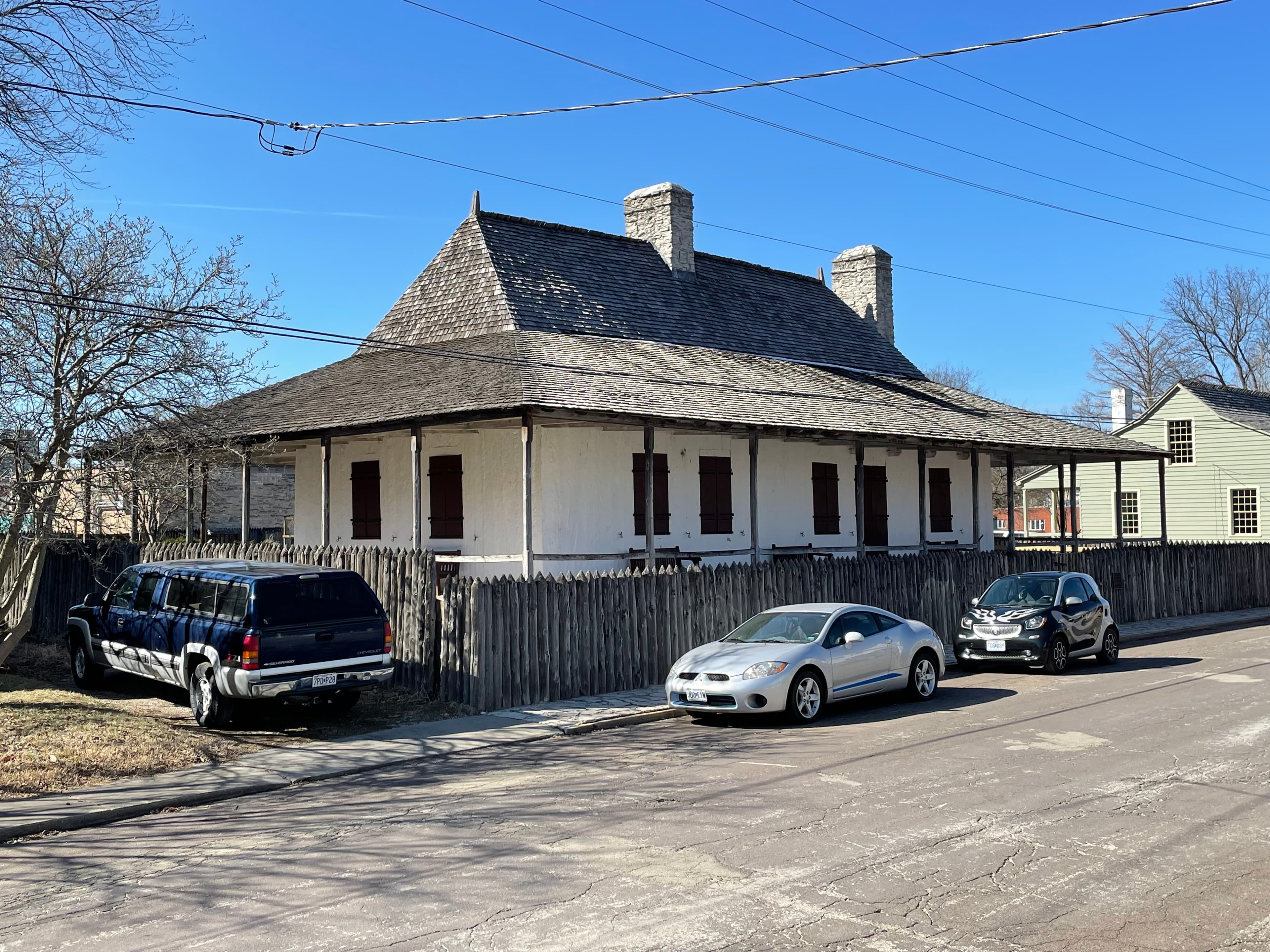 A historic house with a unique roof design surrounded by parked cars and a wooden fence under a clear blue sky.
