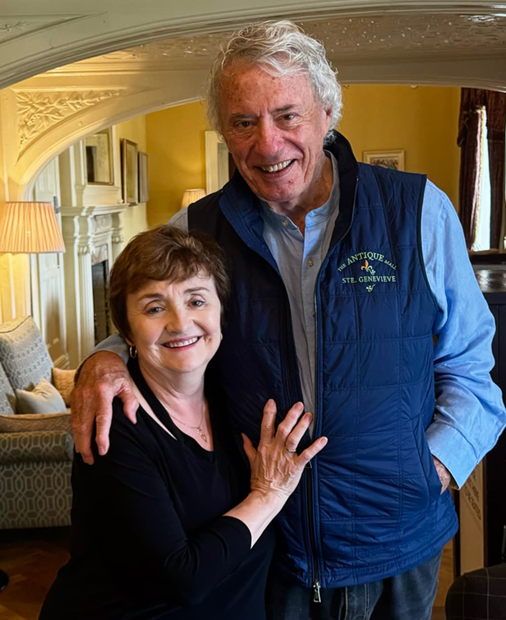 A smiling woman and man pose together with their arms around each other in a cozy interior setting.