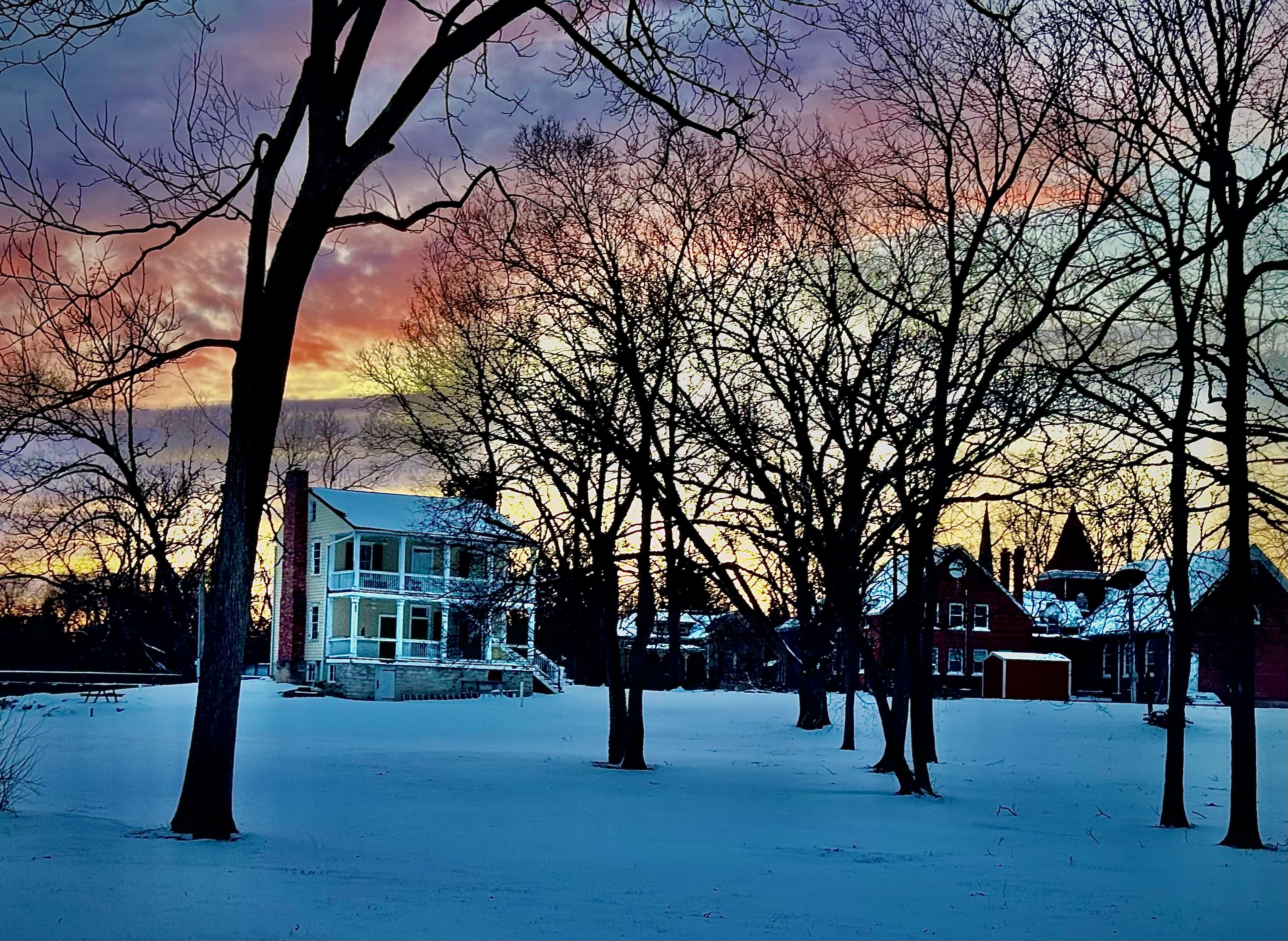 A snow-covered landscape features a vintage house amid bare trees against a colorful sunset sky.